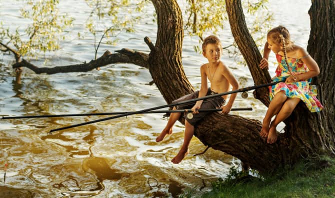 Two children sitting on a tree branch extending over shallow water, with trees and a beach visible in the background.