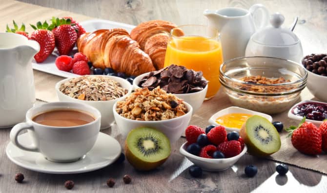 Breakfast spread with coffee, croissants, fresh fruits including strawberries and kiwi, cereal, juice, and pastries on a white table.