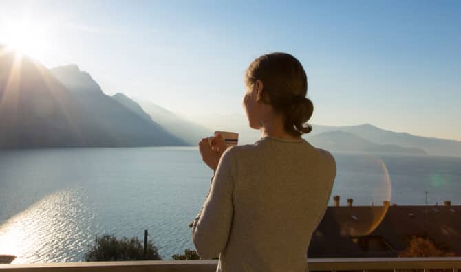 Person with a cup overlooking a scenic mountain lake at sunrise, with dramatic peaks and golden light reflecting on the water.