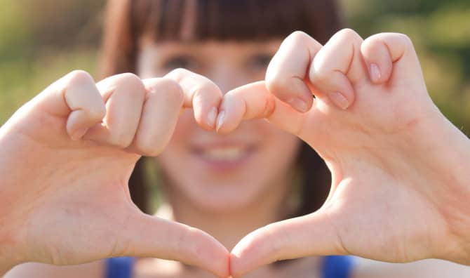 Two hands forming a heart shape with fingers, with a person's smiling face blurred in the background.