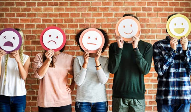 Five people standing against a brick wall, each holding a circular sign with a different emoticon face in front of their heads — from left to right: angry (purple), sad (red/pink), neutral (white), happy (white), and laughing (yellow).