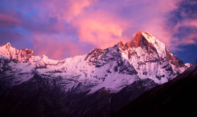 Snow-capped mountain peaks illuminated by pink and purple sunset light with dramatic clouds in the sky.