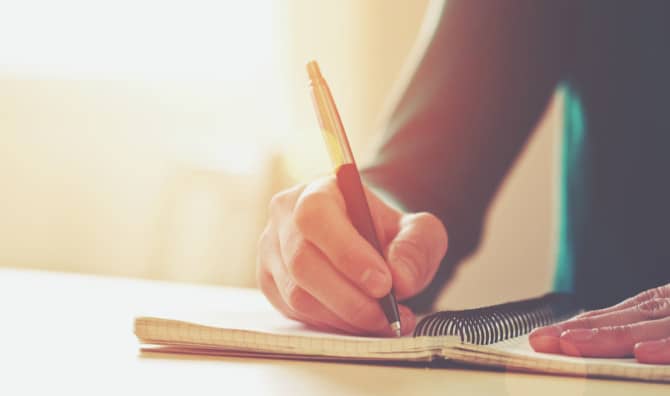 A close-up of a person's hand holding a pen while writing in an open notebook on a wooden desk, bathed in warm, soft natural light from a nearby window.