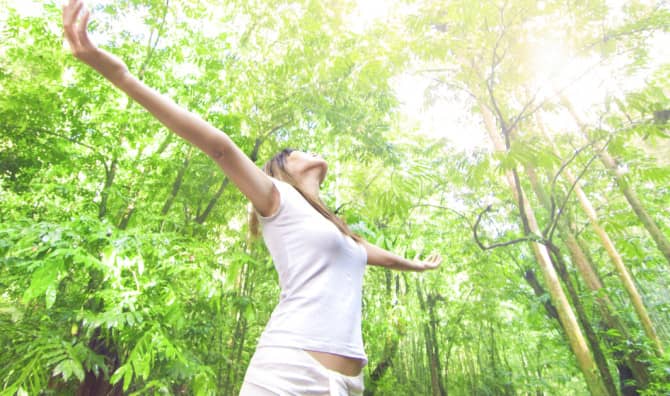 A woman in a white tank top standing in a lush green forest with her arms outstretched and head tilted back, embracing nature as sunlight filters through the leafy tree canopy above.