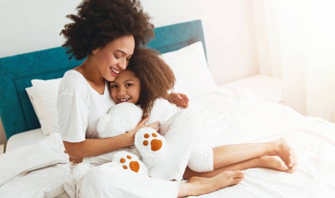 Mother and young child hugging and smiling in bed together, both wearing white with a turquoise headboard behind them.