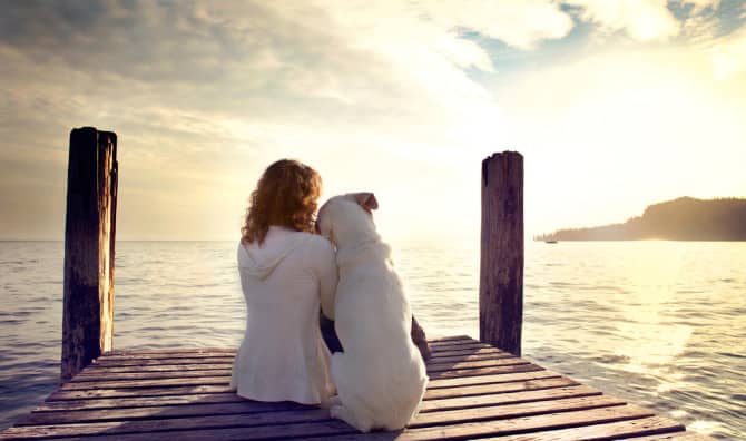Person in a cream sweater sitting on a wooden dock gazing out at calm water at golden hour with mountains visible across the water.
