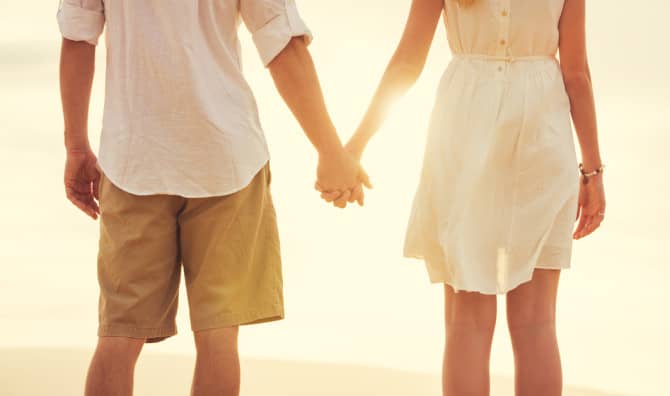 Couple holding hands from behind in casual summer clothing at golden hour with warm backlighting.