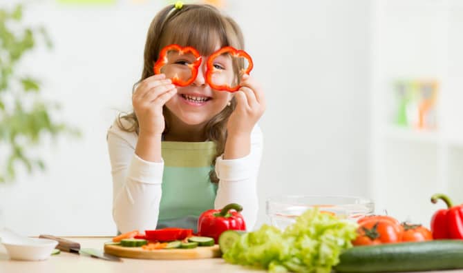 Child holding red bell peppers up to their eyes like glasses and smiling, with fresh vegetables on a cutting board in front of them.
