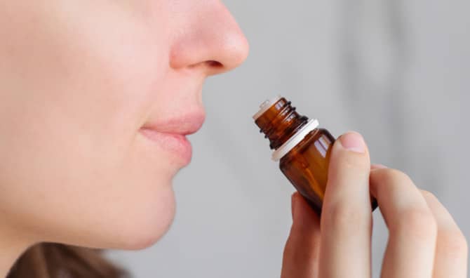 Close-up of a woman smelling a small brown essential oil bottle held near her nose against a gray background.