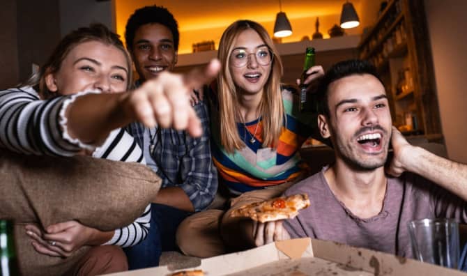 Group of friends gathered indoors pointing at the camera and smiling in warm ambient lighting during a social gathering.