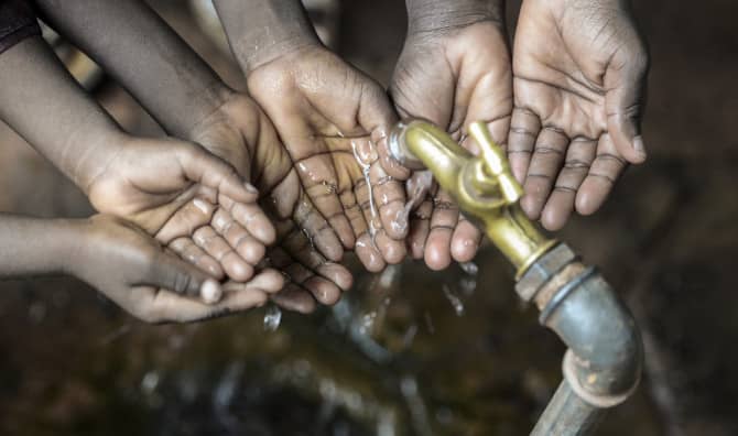 Multiple children's hands cupped together beneath a brass water tap, catching flowing clean water — a symbol of access to safe drinking water.