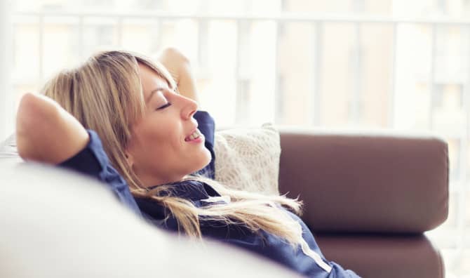 A woman relaxes on a sofa with her hands behind her head, eyes closed, smiling contentedly.