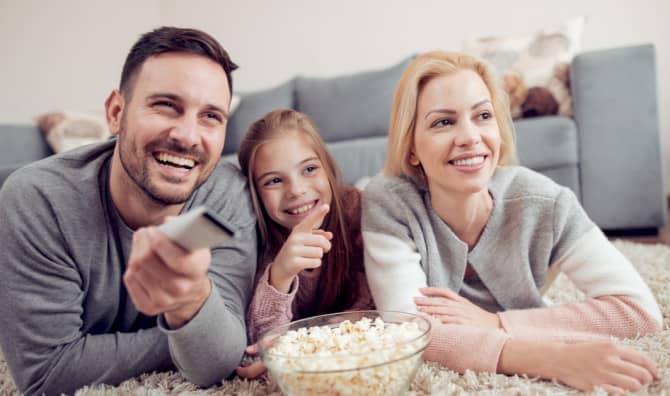 Family of three lying on a couch together eating popcorn and watching a movie, smiling happily.