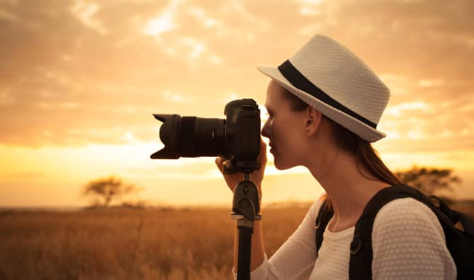 Woman photographer wearing a white straw hat with a backpack, looking through a telephoto lens camera on a tripod aimed at a golden savanna landscape at sunset.