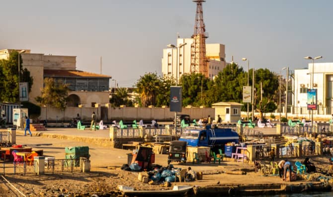 Busy waterfront industrial area with workers, vehicles, equipment, and buildings along a river bank at daytime.