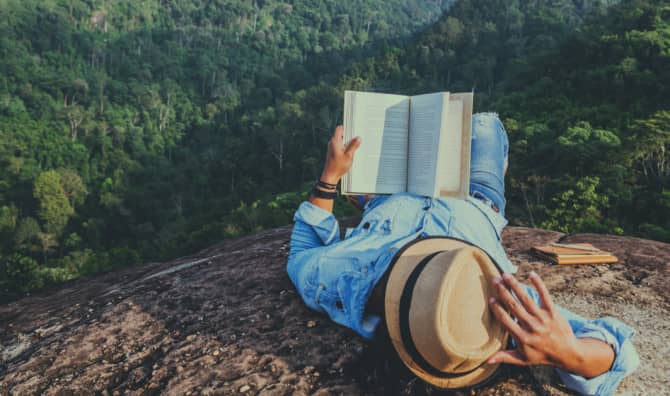 Person lying on a rocky cliff overlooking a dense green forest, reading an open book while holding a straw hat, wearing a blue denim shirt.
