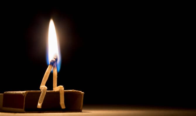 Single candle flame burning on wooden surface against black background, creating warm light in darkness.