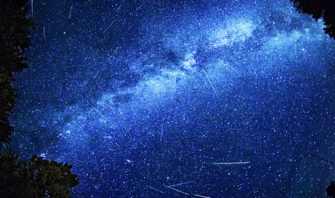 A stunning night sky photograph showing the Milky Way galaxy stretching diagonally across the frame in brilliant shades of blue and white, with numerous shooting stars or meteor streaks visible against the star-filled sky, framed by dark silhouettes of trees in the corners.