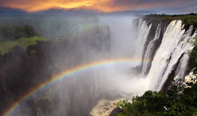 Large waterfall with rainbow in mist, surrounded by lush vegetation under dramatic cloudy sky.