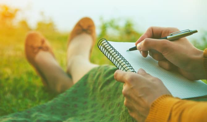 A person sitting on grass in warm sunlight, writing in a spiral-bound notebook resting on their lap, wearing a mustard yellow sweater, green skirt, and tan ballet flats with their legs stretched out.