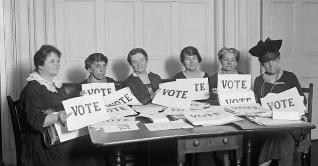A black and white historical photograph of six women seated at a table, each holding signs that read 'VOTE,' advocating for women's suffrage