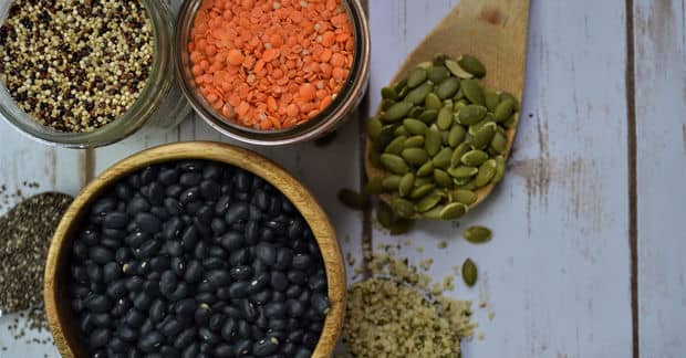 Assortment of plant-based proteins on a white wooden surface: a wooden bowl of black beans, glass jars containing red lentils and tricolor quinoa, a wooden scoop filled with green pumpkin seeds, and scattered hemp hearts.