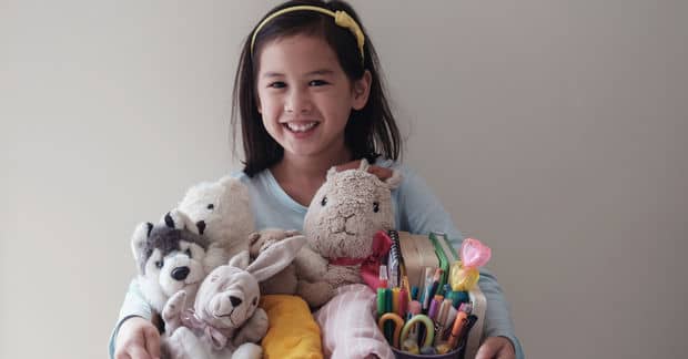 A smiling young girl with a yellow headband holds an armful of stuffed animals, toys, and colorful markers.