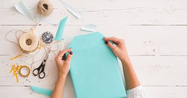Hands holding turquoise cardstock on a white wooden surface surrounded by craft supplies including scissors, twine, and decorative materials.