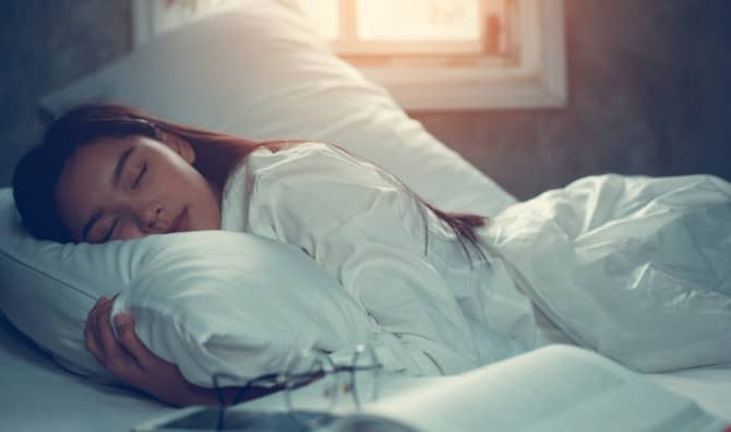 A woman sleeps peacefully on white bedding with a book and glasses nearby as soft light enters through a window.