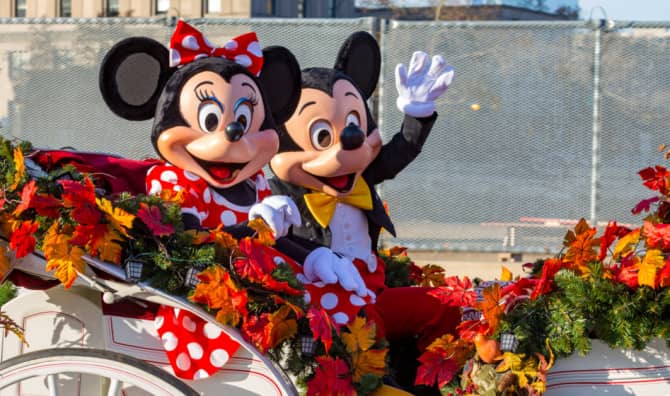 Two costumed theme park characters wave from an autumn-decorated parade float adorned with colorful fall leaves and garlands.