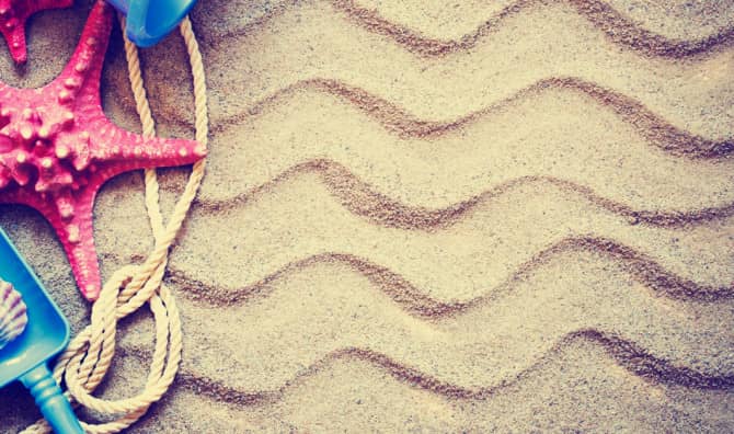 A pink starfish, small seashell, rope, and blue toy shovel on wavy sand, evoking a summer beach theme.