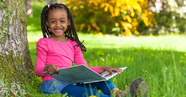 Young girl sitting on the grass by the tree reading a book