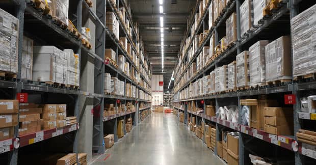 A long warehouse aisle with tall metal shelving units stacked with cardboard boxes and pallets under bright industrial lights.