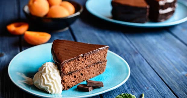 A slice of rich chocolate cake with glossy ganache on a blue plate, served with whipped cream and fresh apricots nearby.