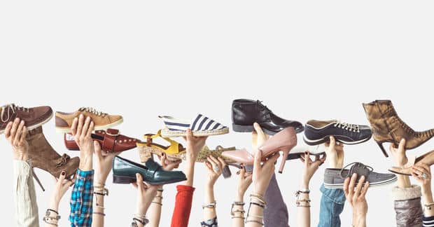 Multiple hands raised in the air, each holding a different style of shoe, against a light gray background.