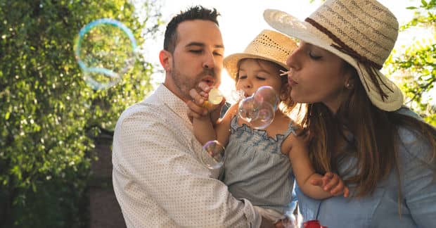 A family of three blows soap bubbles together outdoors on a sunny day, with trees and golden light behind them.