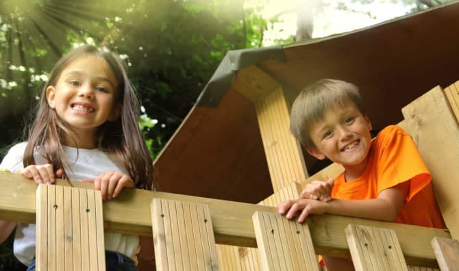 Smiling children look out from a wooden treehouse.