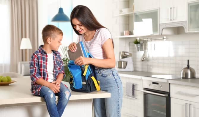 A smiling mother helps her son pack a backpack.