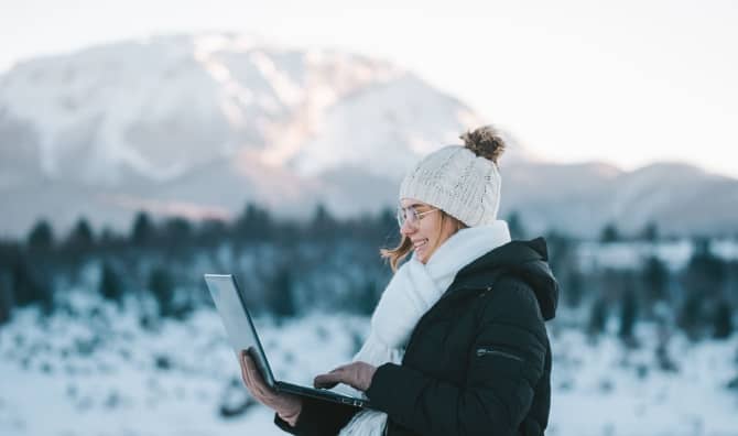 A young smiling woman uses a laptop outdoors in the mountains.