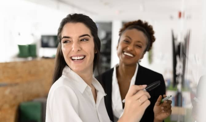Two female coworkers smiling as they prepare to write on a whiteboard.