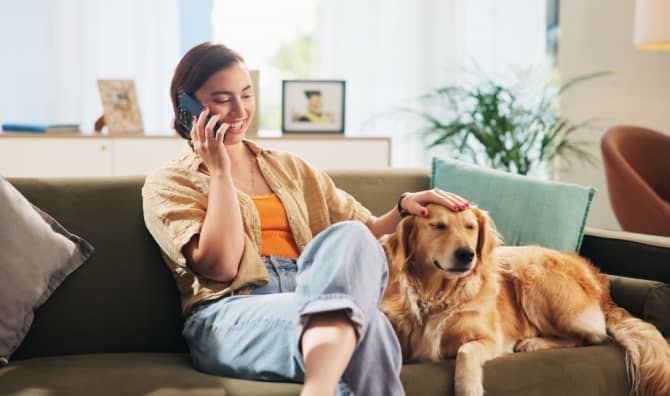 A woman sits on a sofa petting a dog while talking on the phone.