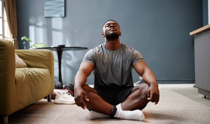 A young adult man sits cross-legged on a carpet in a living room.