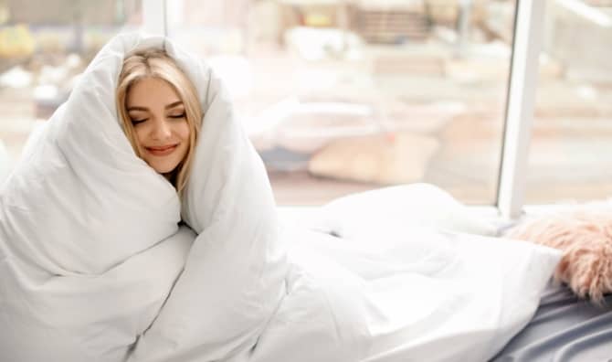 A woman wrapped in a soft blanket sits near a window.