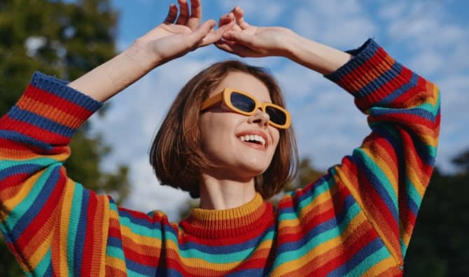 A smiling woman in her twenties wearing a colorful striped sweater and yellow sunglasses.