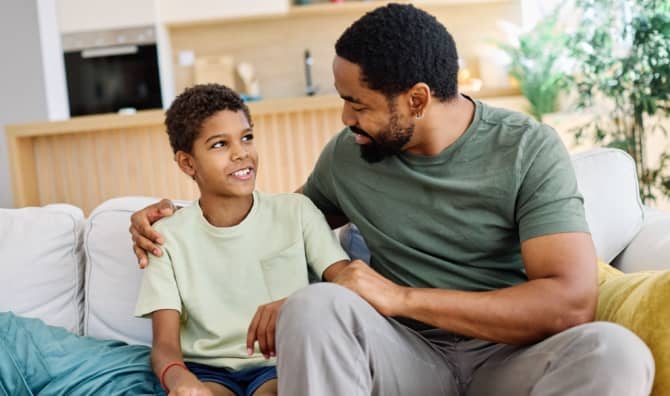 A father and son sit together on a couch at home, talking and smiling.