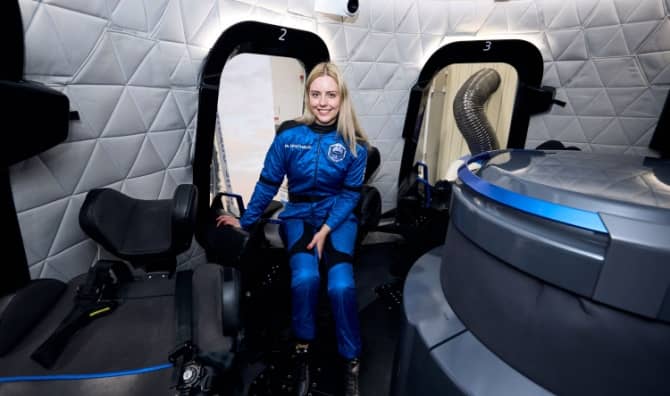 A woman in a blue flight suit sitting inside a spaceflight training capsule.