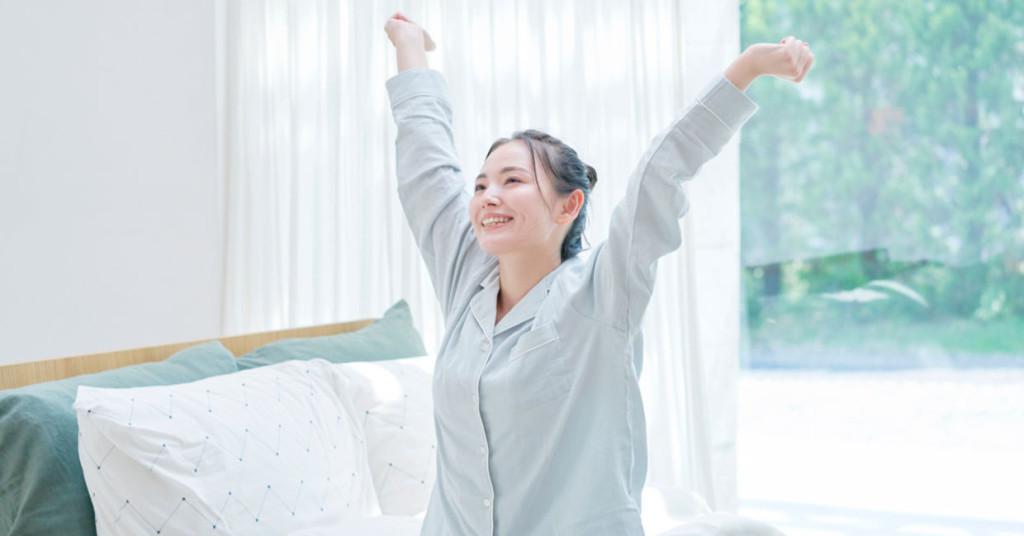 Woman in a light gray shirt stretching with arms raised above her head in a bright bedroom with white curtains and a window overlooking trees