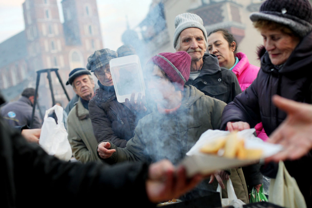Group of people gathering for hot food distribution on a cold urban street, with steam rising and buildings visible in the background.