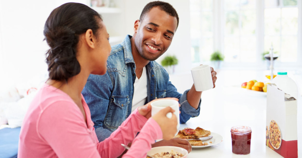 Man and woman smiling while holding mugs during breakfast at a bright kitchen table with food and fruit
