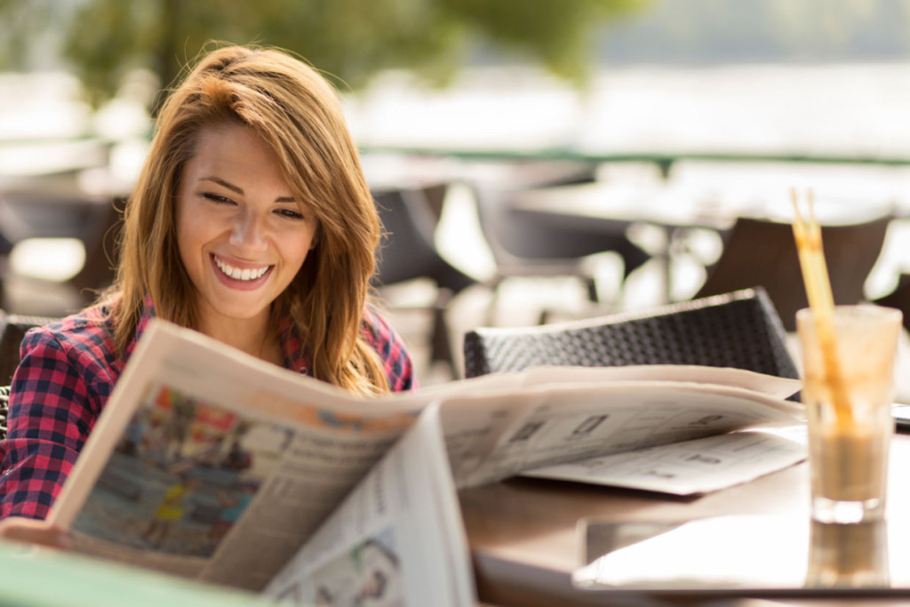 Woman in a pink plaid shirt smiling while reading a newspaper at an outdoor café, with a glass of juice on the table.